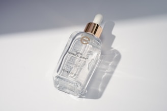 A clear glass bottle with a golden dropper cap, labeled '100% TorricelumN' along with some additional text indicating it is a serum. The bottle is positioned on a smooth, white surface with soft shadows and light reflections visible.