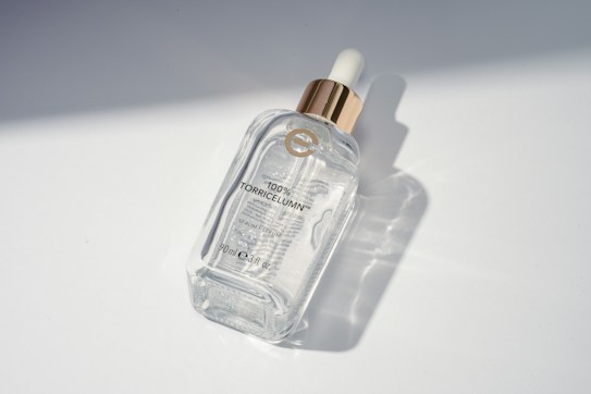 A clear glass bottle with a golden dropper cap, labeled '100% TorricelumN' along with some additional text indicating it is a serum. The bottle is positioned on a smooth, white surface with soft shadows and light reflections visible.