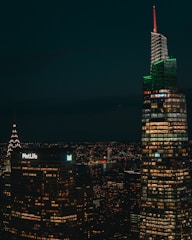 A nighttime cityscape featuring illuminated skyscrapers with numerous windows lit up, including a building with a 'MetLife' sign and another with a distinctive red and green lighting. A river is visible in the background, reflecting city lights.