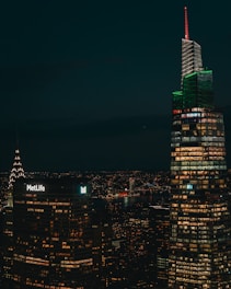 A nighttime cityscape featuring illuminated skyscrapers with numerous windows lit up, including a building with a 'MetLife' sign and another with a distinctive red and green lighting. A river is visible in the background, reflecting city lights.