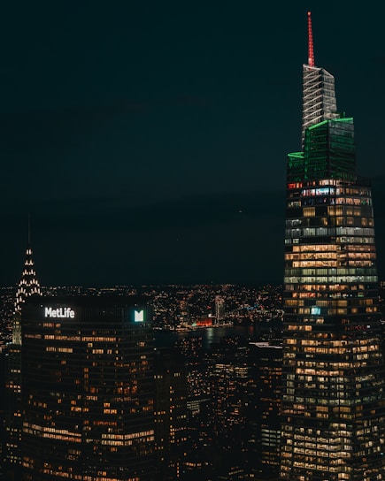 A nighttime cityscape featuring illuminated skyscrapers with numerous windows lit up, including a building with a 'MetLife' sign and another with a distinctive red and green lighting. A river is visible in the background, reflecting city lights.