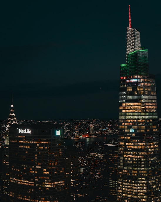A nighttime cityscape featuring illuminated skyscrapers with numerous windows lit up, including a building with a 'MetLife' sign and another with a distinctive red and green lighting. A river is visible in the background, reflecting city lights.