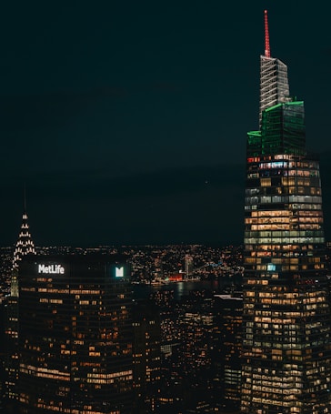 A nighttime cityscape featuring illuminated skyscrapers with numerous windows lit up, including a building with a 'MetLife' sign and another with a distinctive red and green lighting. A river is visible in the background, reflecting city lights.