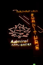 The image features a neon sign glowing in the dark. The sign includes a brightly lit outline of a leaf and the words 'Admiral Appliances' in distinct neon colors—predominantly yellow, white, and red. Another vertical part of the sign spells 'Furniture' from top to bottom in yellow neon. The background is dark, emphasizing the brightness of the neon lights.
