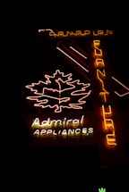 The image features a neon sign glowing in the dark. The sign includes a brightly lit outline of a leaf and the words 'Admiral Appliances' in distinct neon colors—predominantly yellow, white, and red. Another vertical part of the sign spells 'Furniture' from top to bottom in yellow neon. The background is dark, emphasizing the brightness of the neon lights.