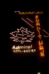 The image features a neon sign glowing in the dark. The sign includes a brightly lit outline of a leaf and the words 'Admiral Appliances' in distinct neon colors—predominantly yellow, white, and red. Another vertical part of the sign spells 'Furniture' from top to bottom in yellow neon. The background is dark, emphasizing the brightness of the neon lights.