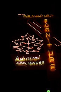 The image features a neon sign glowing in the dark. The sign includes a brightly lit outline of a leaf and the words 'Admiral Appliances' in distinct neon colors—predominantly yellow, white, and red. Another vertical part of the sign spells 'Furniture' from top to bottom in yellow neon. The background is dark, emphasizing the brightness of the neon lights.