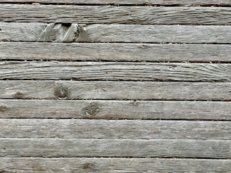 A series of weathered wooden planks aligned horizontally, displaying natural grain patterns and some knot holes. The wood appears aged with a grayish tone, indicating exposure to outdoor elements over time.
