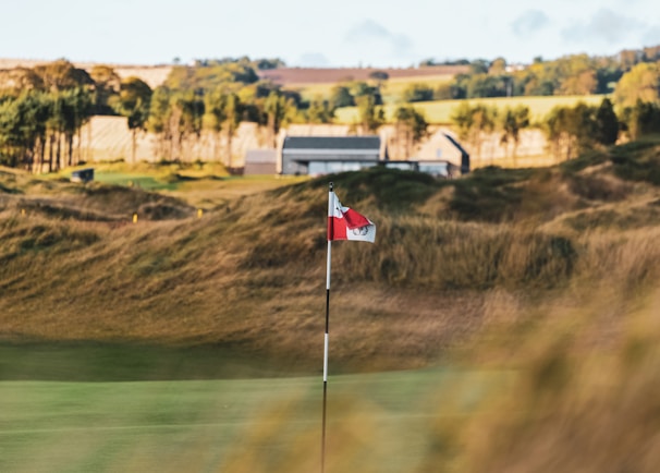 A golf course with a flagstick in the foreground set against a backdrop of rolling hills and a building. The setting appears to be rural with lush greenery and trees lining the horizon.