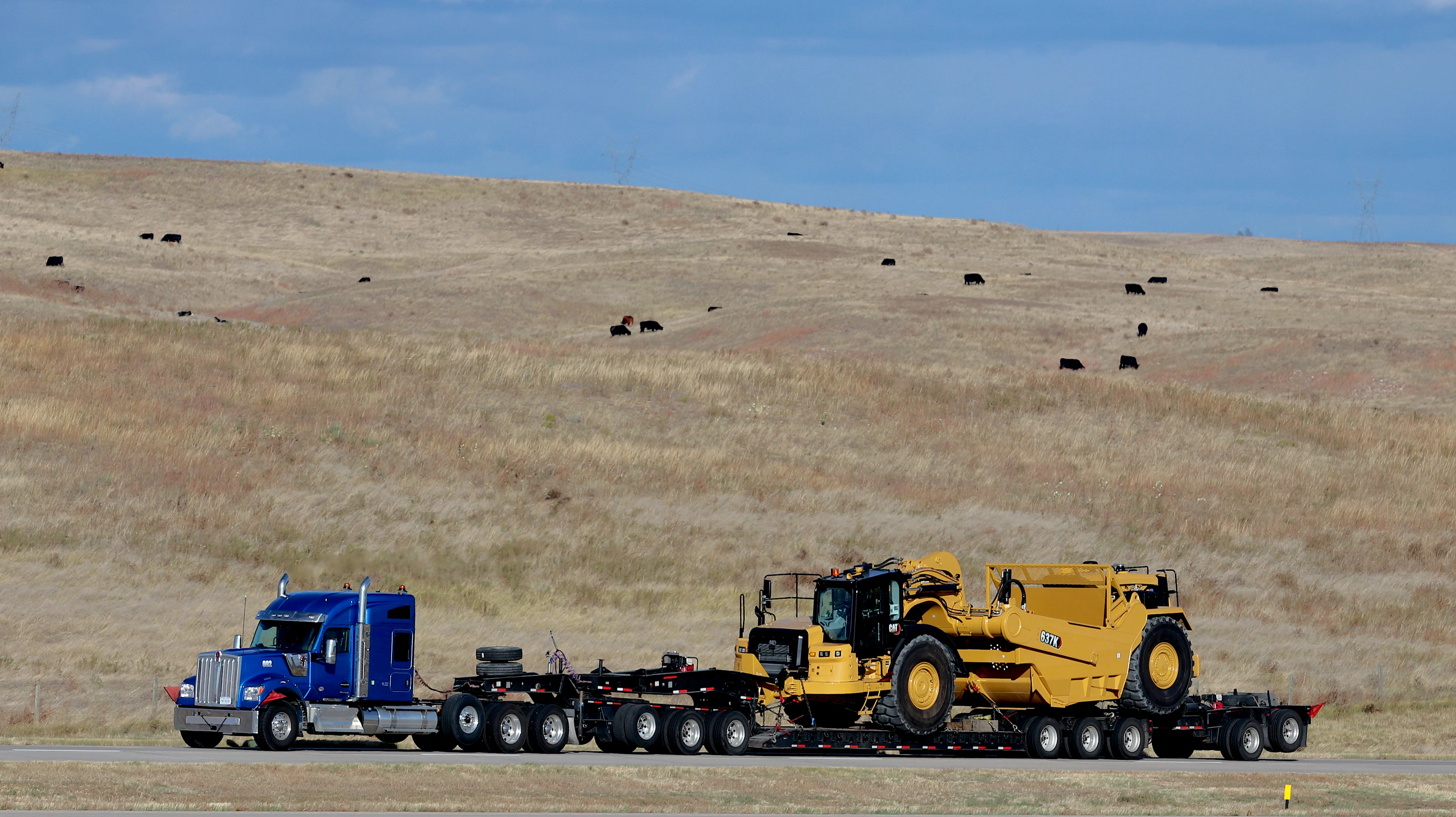 A truck hauling heavy equipment on a lowboy trailer across dry grassland