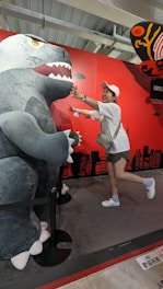 A person playfully interacts with a large dinosaur or monster figure. The figure is gray with sharp teeth, and the setting includes a bright red backdrop with cityscape silhouettes and other decorative elements. The person is wearing a white cap, white shirt, shorts, and sneakers, smiling, and extending their arms toward the figure.