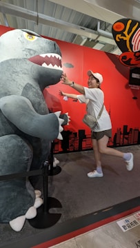 A person playfully interacts with a large dinosaur or monster figure. The figure is gray with sharp teeth, and the setting includes a bright red backdrop with cityscape silhouettes and other decorative elements. The person is wearing a white cap, white shirt, shorts, and sneakers, smiling, and extending their arms toward the figure.