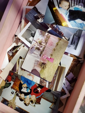 A collection of various printed photographs lies scattered in an open box. The images feature moments such as a baby with a stuffed toy, children playing, and a person in a pink dress near a fence in a backyard. Some photos are in color, while others appear to be black and white. The photos depict a mix of indoor and outdoor scenes.