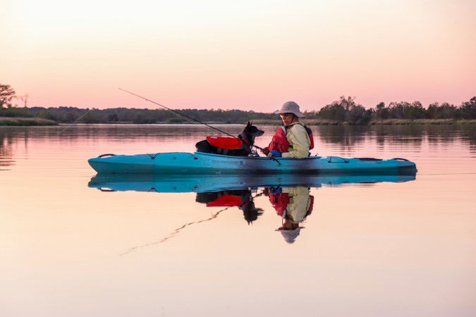 Angler in a sun hat and red life vest fishing from a teal sit-on-top kayak at dusk, a dog in its own red life vest aboard, the pink sky mirrored on still water