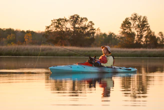 A person wearing a hat is sitting in a light blue kayak on calm water, holding a fishing rod. There is a dog sitting in front of them. The background features trees and grasslands, with the warm glow of the setting or rising sun creating a peaceful scene.