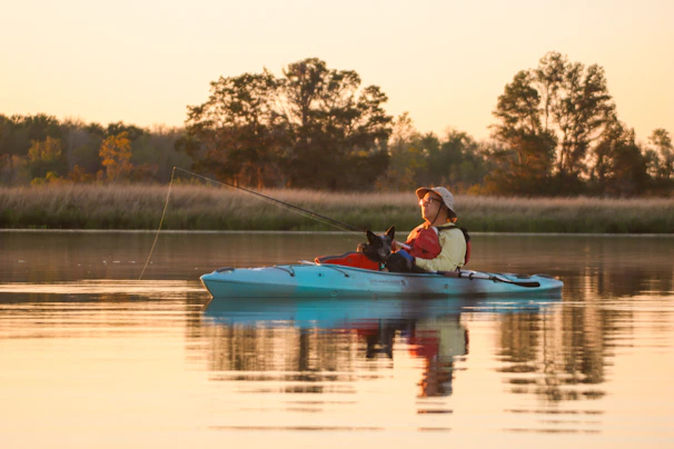 A person wearing a hat is sitting in a light blue kayak on calm water, holding a fishing rod. There is a dog sitting in front of them. The background features trees and grasslands, with the warm glow of the setting or rising sun creating a peaceful scene.