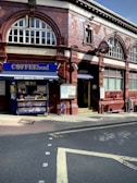 A red-brick building with large arched windows houses an entrance to Hampstead Underground Station, marked by the iconic London Underground sign. Adjacent to the station, a small café called 'COFFEEhead' displays a variety of magazines and snacks.
