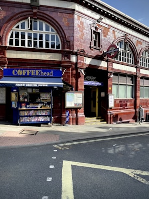 A red-brick building with large arched windows houses an entrance to Hampstead Underground Station, marked by the iconic London Underground sign. Adjacent to the station, a small café called 'COFFEEhead' displays a variety of magazines and snacks.