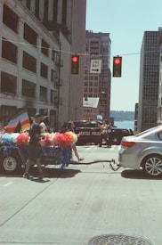 A street scene featuring a decorated float attached to a silver car, with colorful pompoms and a flag, potentially representing LGBTQ+ pride. There are several people involved in the float procession, and the backdrop includes tall buildings and a glimpse of a waterfront. Traffic lights and a 'one way' street sign are visible. A police car labeled 'Seattle Police' is parked across the street.
