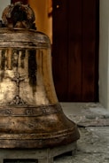 An aged bronze bell with decorative engravings sits on a wooden platform. The bell surface has a patina suggesting age, and it is positioned in front of a partially open wooden door. The background shows part of a stone or concrete floor.