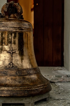 An aged bronze bell with decorative engravings sits on a wooden platform. The bell surface has a patina suggesting age, and it is positioned in front of a partially open wooden door. The background shows part of a stone or concrete floor.