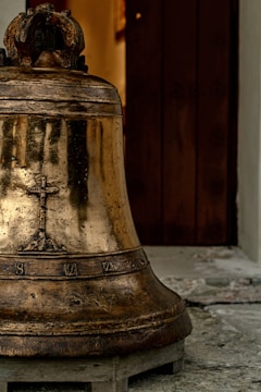 An aged bronze bell with decorative engravings sits on a wooden platform. The bell surface has a patina suggesting age, and it is positioned in front of a partially open wooden door. The background shows part of a stone or concrete floor.