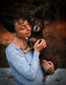 A woman gently holds a small dog in her arms, gazing at it with affection. The setting appears to be outdoors with a blurred background, indicating a natural environment.