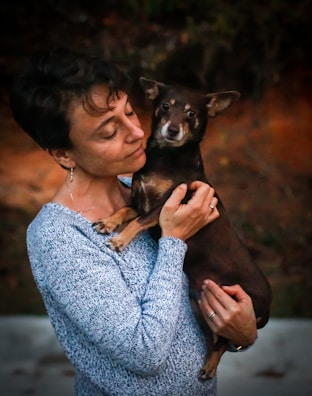 A woman gently holds a small dog in her arms, gazing at it with affection. The setting appears to be outdoors with a blurred background, indicating a natural environment.