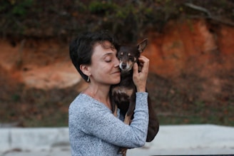 A person gently holding a small dog, with eyes closed and a serene expression, suggesting affection and comfort. The background is a mix of earthy tones and greenery, adding a natural setting.