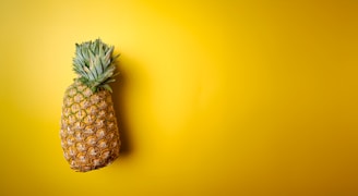 A ripe, whole pineapple with a textured skin and green leafy crown is positioned against a bright yellow background.