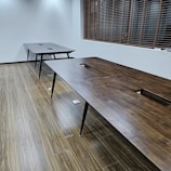 A room with two large, wooden tables positioned parallel to each other on a wood-patterned floor. The tables have black legs and square cutouts in the center. Wooden blinds cover a window on the side wall, and the room has white walls with ceiling lights casting a soft glow.