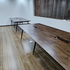 A room with two large, wooden tables positioned parallel to each other on a wood-patterned floor. The tables have black legs and square cutouts in the center. Wooden blinds cover a window on the side wall, and the room has white walls with ceiling lights casting a soft glow.