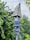 A wooden birdhouse with a pointed, shingled roof painted in shades of blue stands among lush green foliage. A decorative blue ribbon with red and white stars is tied around the base, adding a festive touch to the scene.
