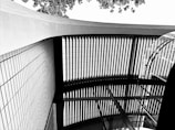 An abstract architectural scene depicting a ceiling with a grid-like structure and a curved glass section. The view includes geometric patterns created by vertical lines and a series of interlocking beams. Above is a bright sky with leaves visible, adding contrast to the structured elements.