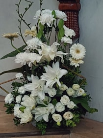 An arrangement of white flowers, including lilies, chrysanthemums, and roses, surrounded by dark green foliage. The display is set against a light-colored wall with a decorative wooden sconce partially visible in the background.