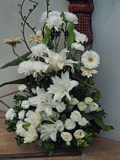 An arrangement of white flowers, including lilies, chrysanthemums, and roses, surrounded by dark green foliage. The display is set against a light-colored wall with a decorative wooden sconce partially visible in the background.