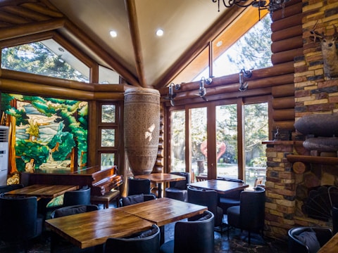 A cozy log cabin interior with wooden furniture and a large textured installation resembling a traditional drum. The room features high ceilings with exposed beams, large windows allowing natural light to flood in, and a colorful stained glass artwork depicting a natural scene with trees and water. There's a stone fireplace with mounted antlers, adding a rustic touch.