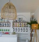 A bar interior with a modern and coastal theme featuring shelves stocked with various bottles of wine and spirits. There are decorative elements like cushions with nautical designs such as anchors, alongside potted plants and a woven lamp. A marble-topped counter displays glasses, a small white statue, and other bar accessories.