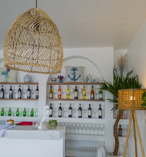 A bar interior with a modern and coastal theme featuring shelves stocked with various bottles of wine and spirits. There are decorative elements like cushions with nautical designs such as anchors, alongside potted plants and a woven lamp. A marble-topped counter displays glasses, a small white statue, and other bar accessories.