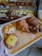 A tray with various freshly baked pastries including croissants and pastries with toppings. In the background, a display case shows an assortment of other baked goods such as rolls and pastries arranged on wooden racks.