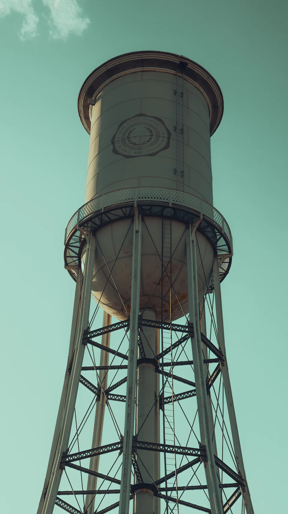 The image shows a tall, cylindrical water tower standing against a clear blue sky with scattered clouds. The tower is painted white and has a black circle on its side. It features two levels, with the top level being slightly smaller than the bottom one. The tower's base is supported by four sturdy metal beams that extend upwards to meet at the top of the structure.