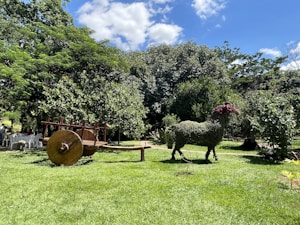 A pastoral scene featuring a large, leafy sculpture of an animal shape next to a rustic wooden cart. The setting is a lush green garden with dense trees and grass under a bright blue sky with scattered clouds.
