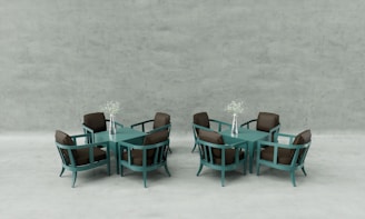 Two sets of modern teal dining tables and chairs, each with four chairs and a central square table. On each table, there is a silver vase holding delicate white flowers. The chairs have brown cushions, and the background is a minimalistic, light grey concrete wall.