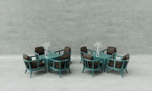 Two sets of modern teal dining tables and chairs, each with four chairs and a central square table. On each table, there is a silver vase holding delicate white flowers. The chairs have brown cushions, and the background is a minimalistic, light grey concrete wall.