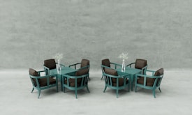 Two sets of modern teal dining tables and chairs, each with four chairs and a central square table. On each table, there is a silver vase holding delicate white flowers. The chairs have brown cushions, and the background is a minimalistic, light grey concrete wall.