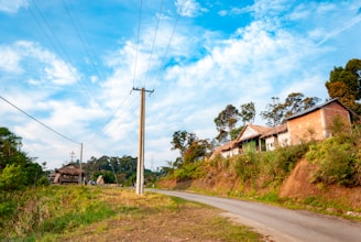 A rural landscape with a narrow paved road curving through a green area. On the right side, there are traditional, rustic houses made of brick and thatch, surrounded by lush vegetation. Tall trees and a few power poles with wires dominate the skyline. The sky is bright with scattered white clouds.