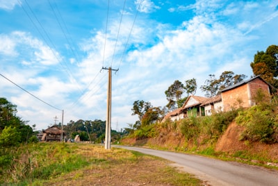 A rural landscape with a narrow paved road curving through a green area. On the right side, there are traditional, rustic houses made of brick and thatch, surrounded by lush vegetation. Tall trees and a few power poles with wires dominate the skyline. The sky is bright with scattered white clouds.