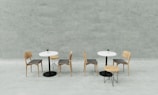 Two small tables with white tabletops are accompanied by four simple wooden chairs and two stools. Each table features a small potted plant as a centerpiece. The setting is minimalistic against a soft gray concrete wall background, creating a clean and modern aesthetic.