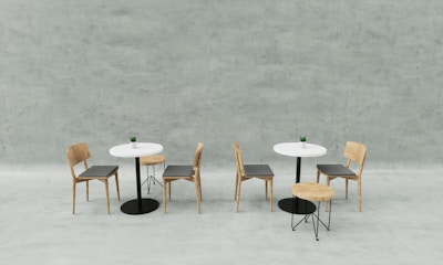 Two small tables with white tabletops are accompanied by four simple wooden chairs and two stools. Each table features a small potted plant as a centerpiece. The setting is minimalistic against a soft gray concrete wall background, creating a clean and modern aesthetic.