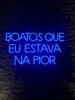 A neon sign displaying a phrase in Portuguese, illuminated in bright blue against a dark brick wall.